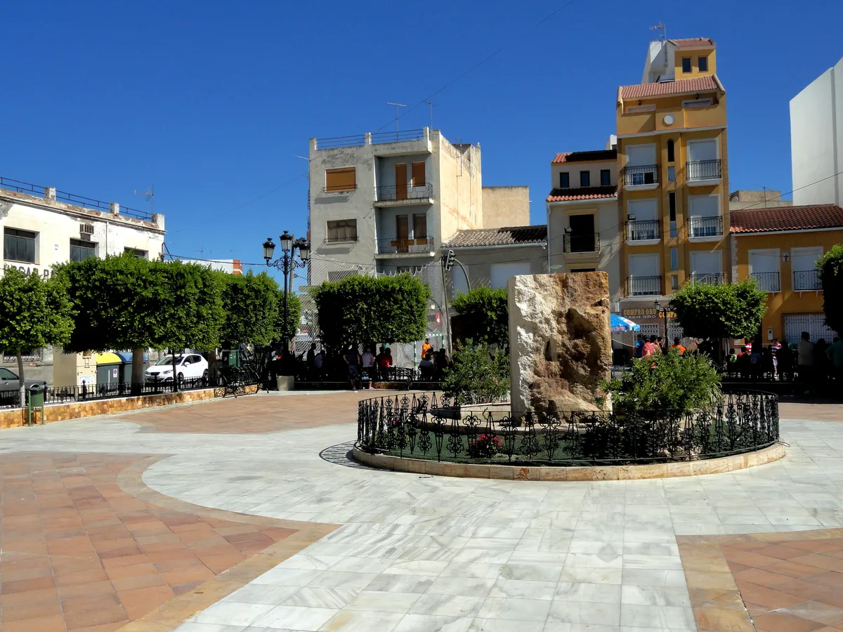 Sunny town square in Albox, Spain, featuring buildings, trees, and a decorative stone monument.