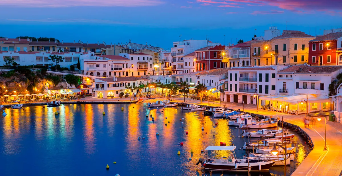 Picturesque harbor town with boats and illuminated buildings reflecting on calm water in Sant Lluis.